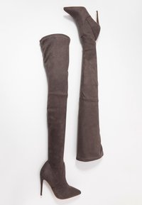 Un paio di stivali alti in suede grigio scuro con tacchi a stiletto e punte affusolate esposti su uno sfondo bianco.