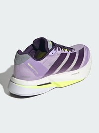 Sapato desportivo roxo com parte superior em rede, detalhes amarelos e três riscas pretas. Apresenta uma sola almofadada e uma marcação proeminente no calcanhar.