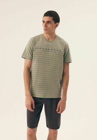 Zielona koszulka w paski z dekoltem w kształcie litery V, z napisem "HENDERSON YACHT CLUB" w granacie. Noszona z ciemnymi szortami. Gładki materiał, luźny krój.