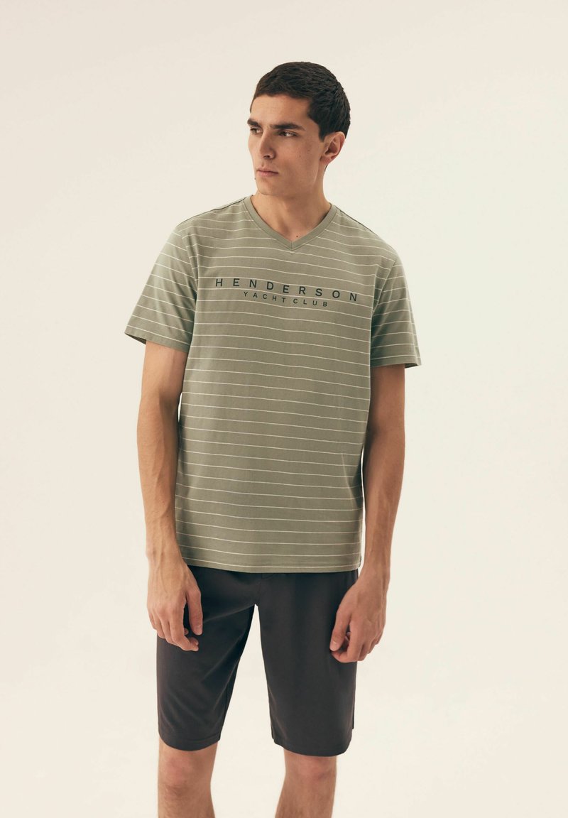 Zielona koszulka w paski z dekoltem w kształcie litery V, z napisem "HENDERSON YACHT CLUB" w granacie. Noszona z ciemnymi szortami. Gładki materiał, luźny krój.