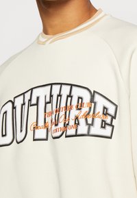 Cream-colored sweatshirt with a round neck, featuring bold black "OUTURE" print and orange text details, made of soft fabric.