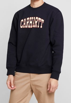 Osoba nosząca czarną bluzę z długim rękawem z napisem "CARHARTT" pogrubionymi białymi i pomarańczowymi literami, w połączeniu z beżowymi spodniami.
