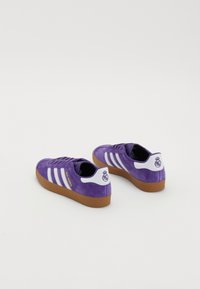 Ténis de camurça roxos com riscas e detalhes brancos. Sola em borracha de goma, acentos de logotipo na parte de trás e texto "Real Madrid" em dourado.