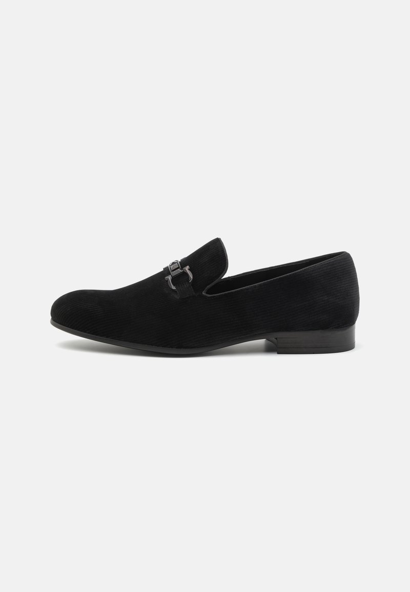 Chaussure slip-on en velours côtelé noir avec un orteil courbé, un accent de boucle en métal et un petit talon, caractérisée par un tissu texturé et un design minimaliste.