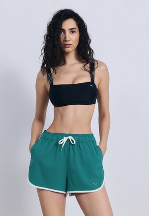 Kvinde med krøllet hår iført sort sports-BH med mærkede stropper og turkise træningsshorts med hvid kant og bindebånd, hænderne i lommerne.