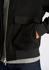 Svart jacka med dragkedja i texturerat tyg och ribbad fåll. Har en sydd framficka och en lager-på-lager grå tröja under.