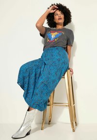 T-shirt graphique avec un design coloré, associé à un pantalon bleu ample à motifs. Bottes de cowboy en métal argenté avec des orteils pointus. Tabouret doré.