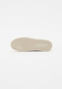 Semelle de chaussure beige avec motif en chevrons, design circulaire près des orteils et marquage de la marque rectangulaire sur surface lisse.