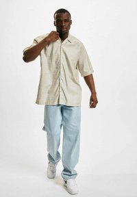 Kortermet beige knappskjorte med et subtile mønster, sammen med lyseblå løse jeans og hvite sneakers med svarte innslag.