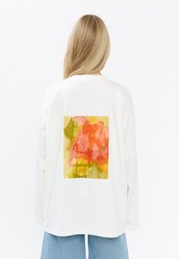 Witte lange mouwen T-shirt met een levendige bloemenprint in roze, geel en groen op de achterkant. Losse pasvorm met verlaagde schouders.