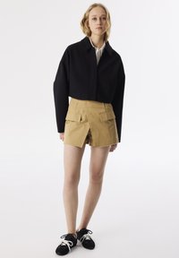 Schwarze Jacke mit Kragen, kombiniert mit beigen Cargo-Shorts mit Fronttaschen und fransigem Saum, getragen mit schwarzen Sneakern.