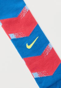 Nike čarape s plavom osnovom i uzorkom crvenih cik-cak pruga, svijetloplavim detaljima i žutim logotipom u sredini dizajna. Pleteni tekstura.