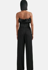 Crni kombinezon bez naramenica s volanima na izrezu, prilagođenim strukom i širokim nogavicama. Stražnji zatvarač i suptilne bočne džepove.