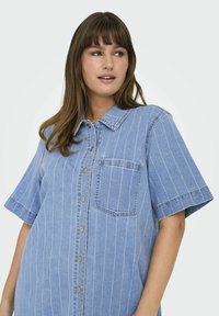 Blå denimskjorta med horisontella vita ränder, korta ärmar, knäppning framtill och bröstficka. Har metallknappar och krage.