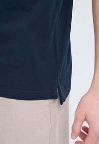 Sötétkék pamut póló texturált felülettel, oldalsó hasítékkal és rövid ujjakkal, bézs nadrággal párosítva. A varrás részletei láthatók.