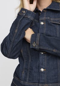 Donkerdenimjack met zichtbare stiksels, metalen knopen en twee borstzakken. De stof heeft een gestructureerde, stevige uitstraling.