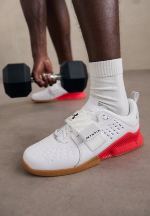 Person hebt schwarze sechseckige Kurzhantel, trägt weiße Sportschuhe mit roten Sohlen und weiße Socken auf einem grauen Boden.