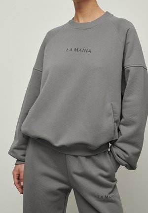 Osoba nosząca szarą bluzę i dopasowane spodnie, z logo "LA MANIA" wydrukowanym dużym, ciemnoszarym tekstem z przodu.