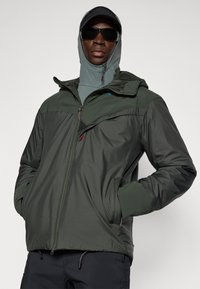 Jachetă zip-up de culoare verde închis, cu glugă, având un guler contrastant de culoare verde deschis, buzunare laterale și o textură netedă, rezistentă la apă.