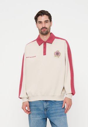 Uomo che indossa una felpa beige e rossa con colletto con testo ed emblema "Fear Nothing", abbinata a jeans blu, in piedi su sfondo bianco.