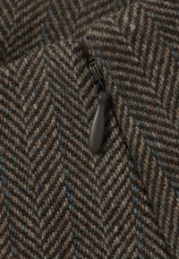 Vlnitá herringbone textília v hnedej farbe s modrými akcentami, s uzáverom na zips. Textúra je hrubá, čo zdôrazňuje zložitý tkaný vzor.