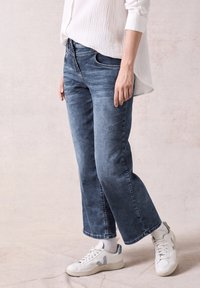 Jeans de mezclilla de pierna ancha en azul claro con una textura desgastada, combinados con zapatillas blancas y detalles grises, llevados con una camisa blanca holgada.