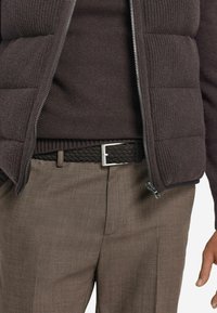 Mannenoutfit met een donkerbruine gestikte vest over een donkere trui, gecombineerd met gestructureerde bruine broeken en een gevlochten leren riem met een zilveren gesp.