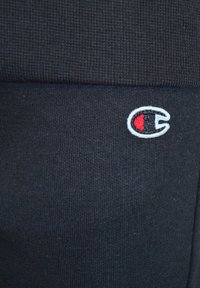 Svart ribbad textil med en liten broderad logotyp med röd accent. Fokusera på materialets textur och logotypdesign.