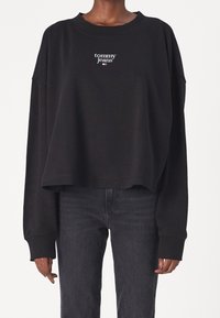 Felpa nera con un design cropped e maniche lunghe. Presenta un logo bianco "tommy jeans" sul petto. Il materiale sembra morbido.