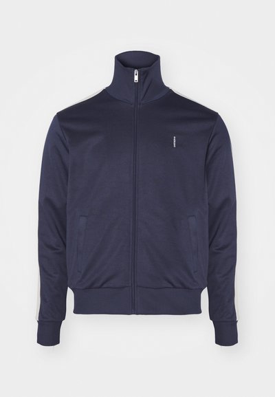 Marinfärgad zip-up jacka med hög krage, två sidofickor och kontrasterande ränder längs ärmarna. Tillverkad av mjukt, slätt tyg.