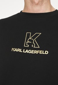 T-shirt di cotone nero con una grafica dorata della lettera "K" e una silhouette profilata, con il testo "KARL LAGERFELD" sottostante.