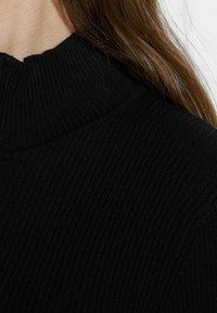 Pull en maille côtelée noir avec un col montant. Le tissu a une texture avec des lignes verticales, offrant une apparence ajustée et près du corps.