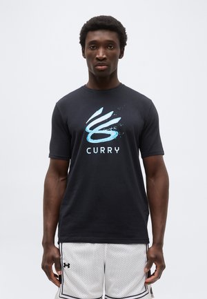 Νεαρός άνδρας που φοράει μαύρο μπλουζάκι με το λογότυπο Curry και λευκά μπουφάν μπάσκετ Under Armour, στέκεται μπροστά σε απλό λευκό φόντο.