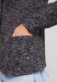 Main dans la poche d'une veste en laine à carreaux gris et noirs avec un petit détail d'étoile argentée, portée sur un jean en denim bleu clair.