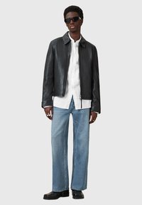 Giubbotto di pelle nera con colletto, abbinato a una camicia bianca e jeans larghi azzurri. Gli occhiali da sole neri completano il look.