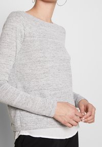 Suéter gris claro de manga larga y cuello redondo. Presenta un patrón de punto texturizado y un dobladillo en capas con un borde blanco.