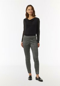 Czarna bluzka z długim rękawem, luźno upięta, zestawiona z szarymi spodniami o dopasowanym kroju w kratę. Model nosi błyszczące czarne mokasyny. Prosty, nowoczesny design.