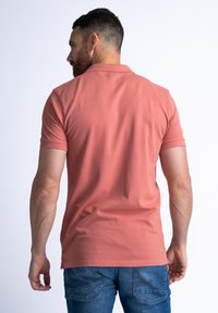 Polo shirt pour homme à manches courtes en rose pâle, fabriqué en coton. Il présente un col classique et une coupe décontractée avec des fentes sur les côtés à l'ourlet.