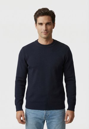 Uomo con capelli corti castani che indossa un maglione blu navy con bottoni sulle spalle e jeans azzurri, in piedi davanti a uno sfondo semplice.