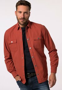 Rotes Baumwollhemd mit langen Ärmeln, zwei Brusttaschen und silbernen Druckknöpfen. Über einem dunklen Oberteil getragen, kombiniert mit Jeans.