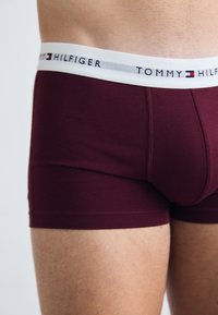 Bordeauxfarbene Baumwollboxershorts mit einem weißen elastischen Bund, der mit dem Text und Logo von "TOMMY HILFIGER" versehen ist. Glatte Textur, enganliegende Passform.