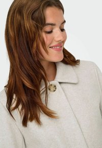Giovane donna con lunghi capelli castano-rossastri sorride dolcemente, indossando un cappotto beige chiaro con un grande bottone vicino al colletto.
