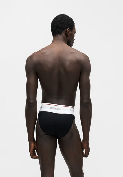 Άνδρας με σκούρο δέρμα και κοντά σγουρά μαλλιά, που στέκεται με την πλάτη προς τα εμπρός, φοράει μαύρα εσώρουχα με λευκή μέση με την ένδειξη "DSQ2" σε μονόχρωμο φόντο.