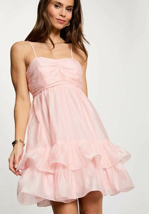 Donna che indossa un abito rosa chiaro senza maniche con volant e spalline sottili, in piedi davanti a uno sfondo chiaro e uniforme.