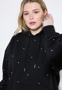 Sudadera negra de tela suave, con embellishments parecidos a perlas dispersos, capucha con cordones y bolsillo tipo canguro.