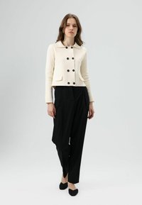 Veste courte de couleur crème avec des boutons noirs, design ajusté, et deux poches avant, associée à un pantalon noir taille haute.