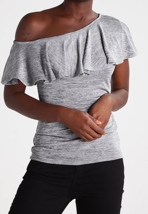 Szary top z opuszczanymi ramionami i falbaną przy dekolcie, wykonany z miękkiej, teksturowanej tkaniny. Do tego dopasowane czarne spodnie, ręce złożone przed sobą.