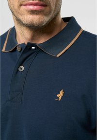 Jūrinė polo marškinėliai su rudais apykaklės akcentais ir maža išsiuvinėta logotipu. turi du mygtukus ir tekstūruotą audinį.