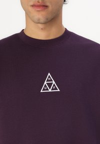 Violetinis džemperis su apvalia iškirpte ir baltu siuvinėtu trikampiu logotipu, kuriame yra "HUF" inicialai krūtinėje. Minkšta medžiagos tekstūra.