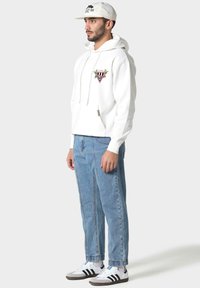 Uomo che indossa una felpa bianca con grafica, jeans azzurri chiari, sneakers bianche e cappellino bianco, in piedi davanti a uno sfondo chiaro e neutro.